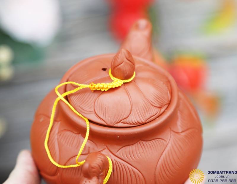 Nắp ấm sen Bát Tràng được tạo hình nụ sen khum tròn, khắc chìm cánh nổi đồng nhất với thân ấm. Dây nối vàng tạo điểm nhấn thẩm mỹ, giữ nắp chắc chắn và mang ý nghĩa tài lộc, bền chặt.