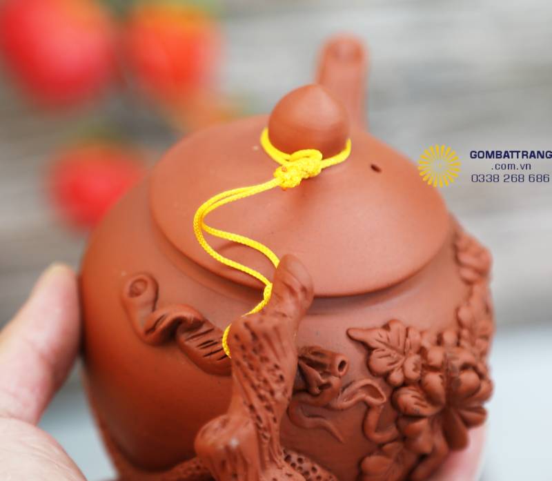 Cận cảnh phần nắp và quai ấm, nơi người thợ gốm tỉ mỉ khắc từng cánh hoa. Sắc đỏ đất hòa với dây vàng, giản dị mà nổi bật, gợi cảm giác ấm áp như nắng sớm.