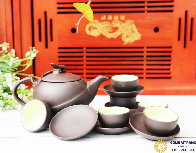 Cận cảnh bộ trà đặt trên nền gỗ đỏ, nhấn mạnh vào tông màu nâu đen của thân ấm và ánh men xanh non trong lòng chén. Dáng ấm tròn gọn, vòi rót cong tự nhiên, nắp khít vừa, đảm bảo giữ nhiệt tốt.