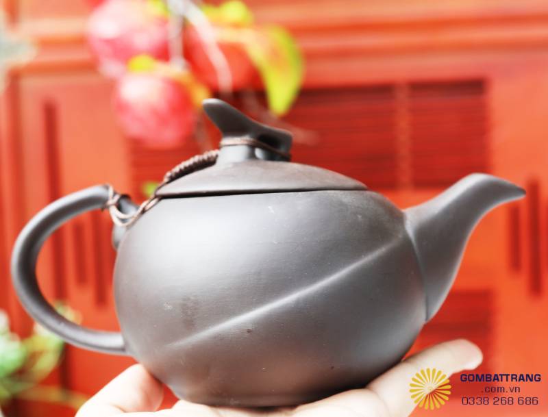 Góc chụp thẳng từ phía trước ấm trà, thể hiện sự cân đối giữa vòi, thân và quai cầm. Dáng ấm tròn hài hòa, dây da buộc nắp tạo điểm nhấn thủ công. Sản phẩm thể hiện độ tinh xảo trong căn chỉnh tỷ lệ.