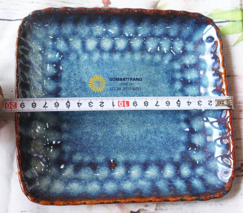 chiếc đĩa vuông bản lớn với thước đo đặt ngang (khoảng 20-22cm). Bề mặt đĩa có các vân lõm nhẹ giúp thoát nước hoặc tạo điểm nhấn cho món ăn, viền nâu hổ phách nổi bật trên nền xanh hỏa biến.